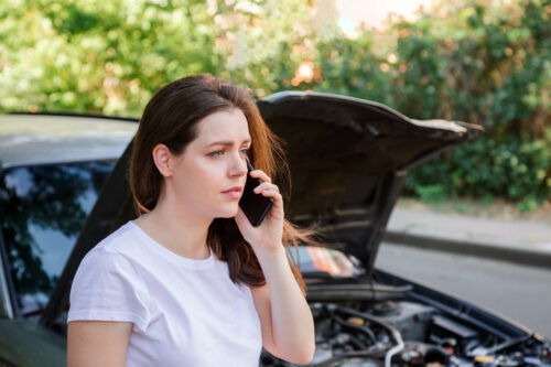 Portrait of frustrated young woman calling mobile phone for help Insurance service or ambulance after car accident. Woman in front of broken car with a mobile phone in hand.