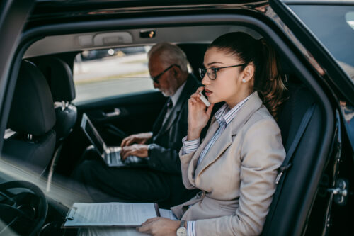 Good looking senior business man and his young woman colleague or coworker sitting on backseat in luxury car. They talking, smiling and using laptop and smart phones. Transportation in corporate business concept.