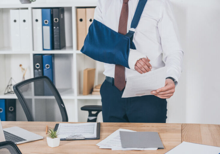 cropped view of worker with broken arm in bandage standing and holding paper over table in office, compensation concept