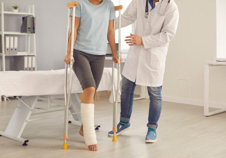 Patient with broken leg assisted by doctor. Injured young woman stands up from examination bed at hospital and walks using crutches with help of medical specialist. Rehabilitation after injury concept